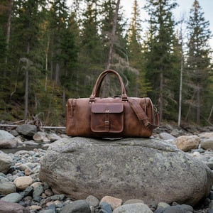 May include: A brown leather duffel bag featuring a rectangular front pocket with a buckle closure, two top handles, and a detachable shoulder strap. The bag has a structured design with visible stitching and metal hardware accents on the exterior.