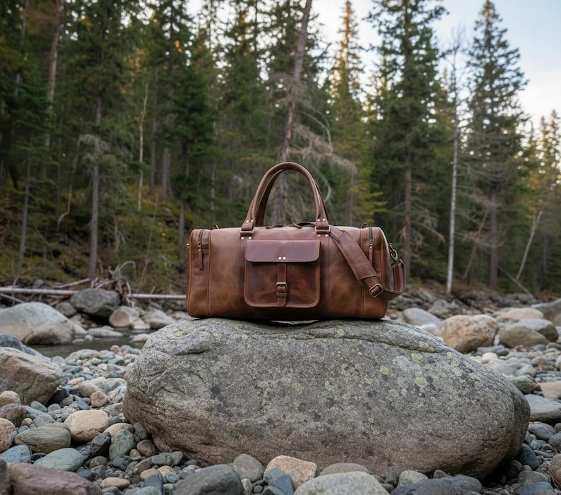 May include: A brown leather duffel bag featuring a rectangular front pocket with a buckle closure, two top handles, and a detachable shoulder strap. The bag has a structured design with visible stitching and metal hardware accents on the exterior.