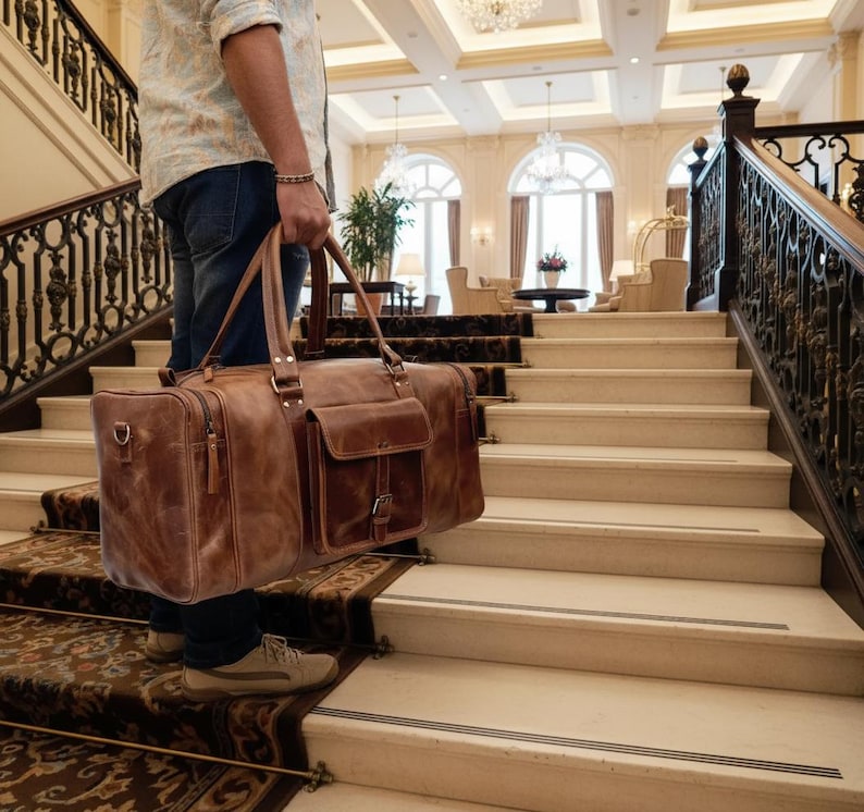 May include: A large brown leather duffel bag with two top handles, a rectangular front pocket with a buckle closure, and side zippers. The bag features visible stitching, metal hardware, and a structured rectangular shape for travel storage.