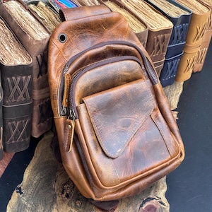 Könnte beinhalten: Eine braune Leder-Umhängetasche mit einer Vordertasche und einem Reißverschluss-Hauptfach. Die Tasche hat ein abgenutztes, abgenutztes Aussehen. Die Tasche wird neben einer Sammlung alter Bücher präsentiert.