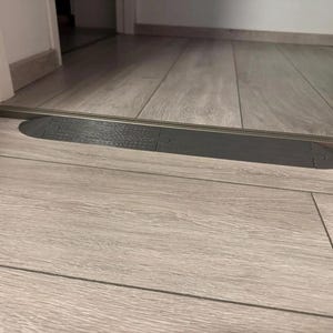May include: A dark gray threshold ramp installed between two rooms. The ramp is made of a textured material and is designed to provide a smooth transition between different floor levels. The floor is light gray wood.
