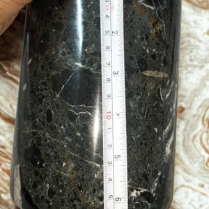 May include: A cylindrical, dark green and black marble container with white veining. The container is approximately 4 inches tall. A measuring tape is placed next to the container.