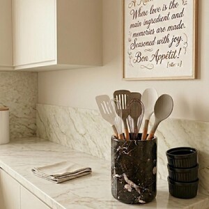 May include: A kitchen scene with a marble countertop and a framed sign that reads "A Kitchen's Heart: Where love is the main ingredient and memories are made. Seasoned with joy. Bon App&eacute;tit!". A black and white marble utensil holder contains various kitchen tools.