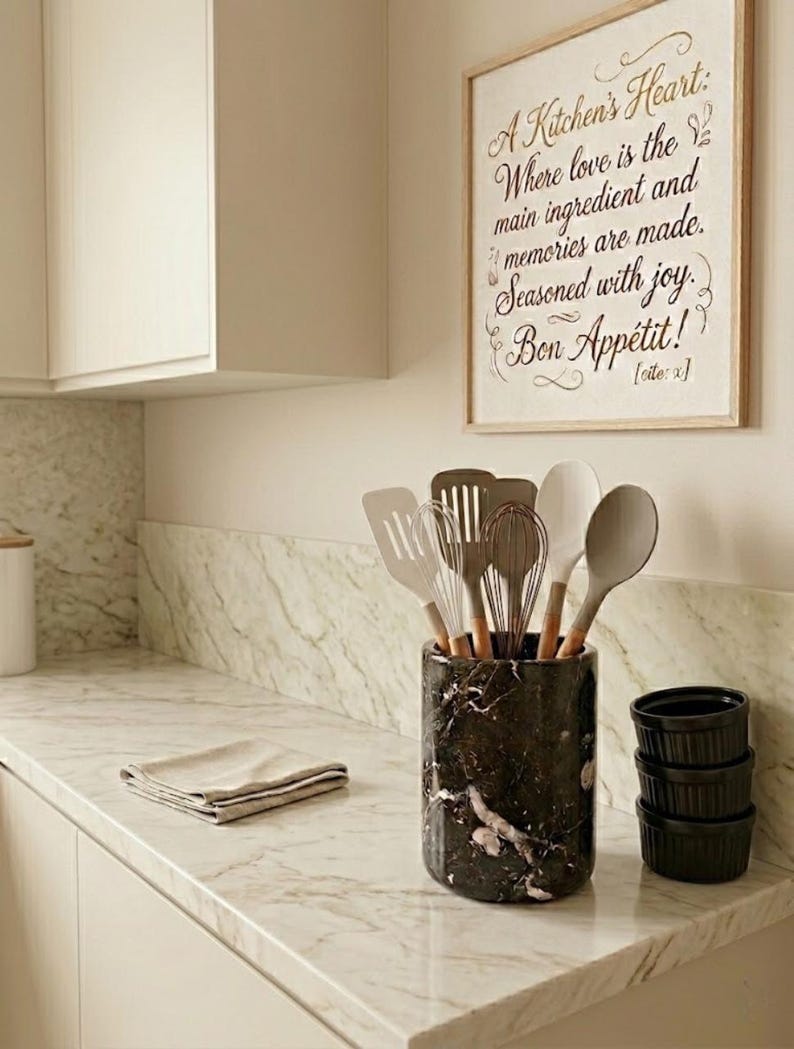 May include: A kitchen scene with a marble countertop and a framed sign that reads "A Kitchen's Heart: Where love is the main ingredient and memories are made. Seasoned with joy. Bon App&eacute;tit!". A black and white marble utensil holder contains various kitchen tools.