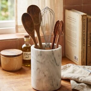 May include: A marble utensil holder with wooden spoons, a whisk, and tongs. A small jar of olive oil and a cork coaster are also visible. The items are arranged on a wooden cutting board, suggesting a kitchen theme.