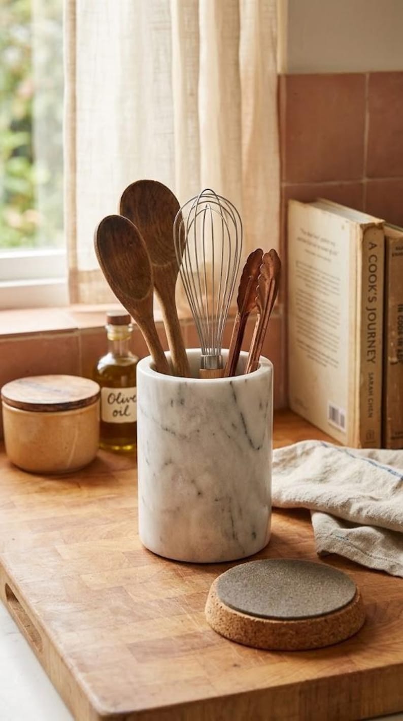 May include: A marble utensil holder with wooden spoons, a whisk, and tongs. A small jar of olive oil and a cork coaster are also visible. The items are arranged on a wooden cutting board, suggesting a kitchen theme.