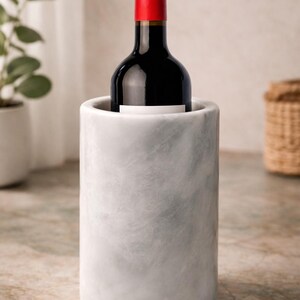 May include: A marble wine chiller holds a bottle of red wine. The wine bottle has a red cap and a white label. The chiller is cylindrical and has a gray and white marbled pattern.
