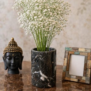 May include: A black marble vase with white veining holds a bouquet of white flowers. A small black Buddha statue with gold accents and a decorative picture frame are also on display.