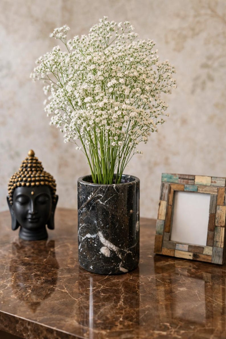 May include: A black marble vase with white veining holds a bouquet of white flowers. A small black Buddha statue with gold accents and a decorative picture frame are also on display.
