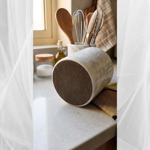 May include: A white marble mortar with gray veining, featuring a dark brown base. Kitchen utensils, including wooden spoons, a whisk, and tongs, are visible in the background. The mortar is on a light gray countertop, suggesting a culinary theme.