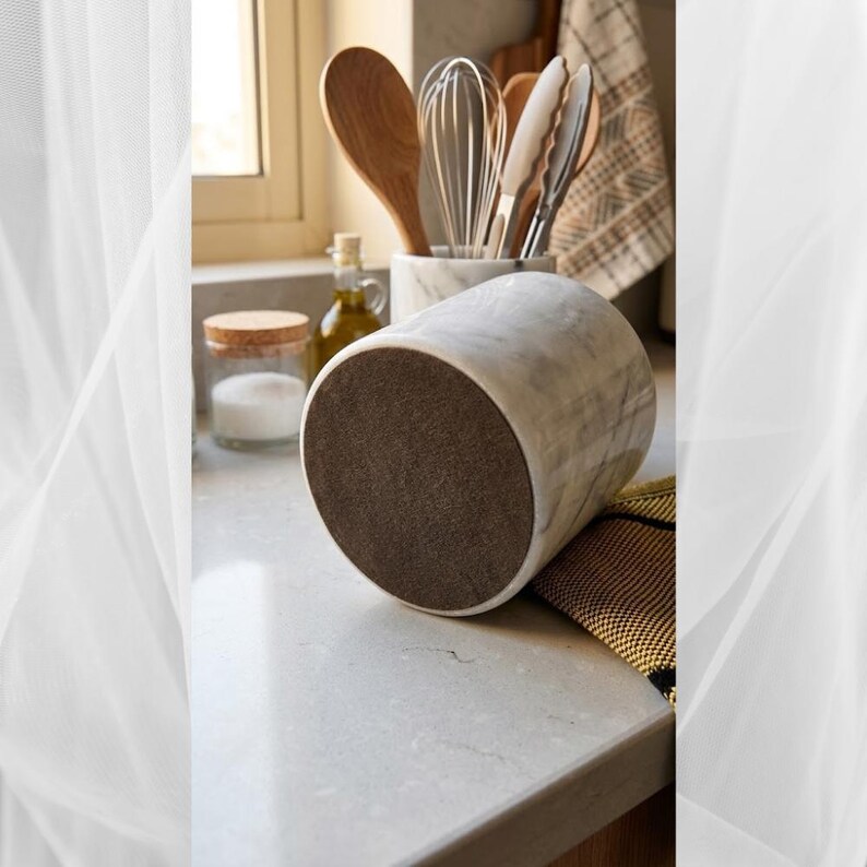 May include: A white marble mortar with gray veining, featuring a dark brown base. Kitchen utensils, including wooden spoons, a whisk, and tongs, are visible in the background. The mortar is on a light gray countertop, suggesting a culinary theme.