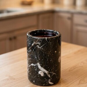 May include: A cylindrical black marble container with white veining, filled with a dark liquid. The container sits on a light wood surface, with a blurred kitchen background.