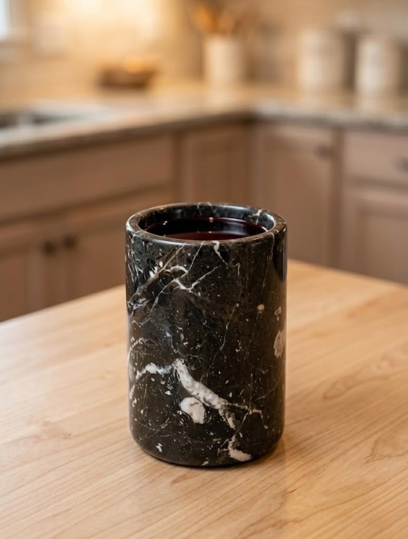 May include: A cylindrical black marble container with white veining, filled with a dark liquid. The container sits on a light wood surface, with a blurred kitchen background.