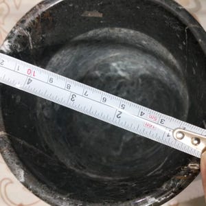 May include: A dark gray mortar with a tape measure showing a diameter of approximately 4 inches. The mortar has a rough, textured surface and a rounded interior. The tape measure is blue and white.