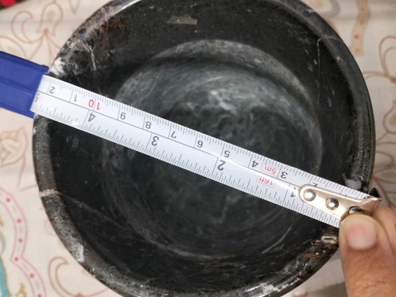 May include: A dark gray mortar with a tape measure showing a diameter of approximately 4 inches. The mortar has a rough, textured surface and a rounded interior. The tape measure is blue and white.