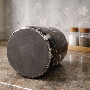 May include: A dark, cylindrical object with a textured, distressed finish, possibly a decorative container or vase. The base is a dark gray, circular shape. The background includes a tray with salt and pepper shakers.