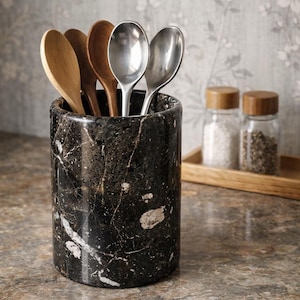 May include: A black marble utensil holder with white veining, filled with wooden and stainless steel spoons. The kitchenware is displayed on a countertop with jars of spices in the background.
