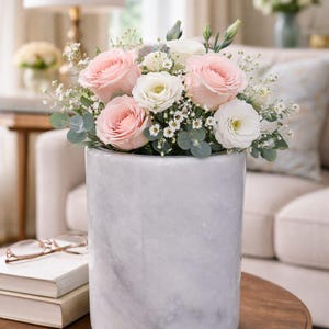 May include: A floral arrangement featuring pink roses, white roses, and greenery in a cylindrical marble vase. The vase sits on a dark wooden table. A lamp and a sofa are visible in the background, creating a soft, inviting ambiance.