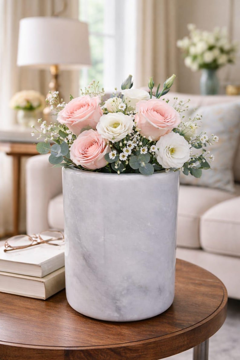 May include: A floral arrangement featuring pink roses, white roses, and greenery in a cylindrical marble vase. The vase sits on a dark wooden table. A lamp and a sofa are visible in the background, creating a soft, inviting ambiance.
