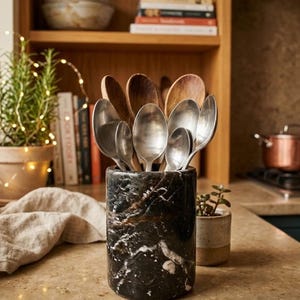 May include: A black and gold marble utensil holder filled with wooden and silver spoons. The cylindrical holder sits on a light-colored countertop. A small potted plant and a stack of books are in the background.