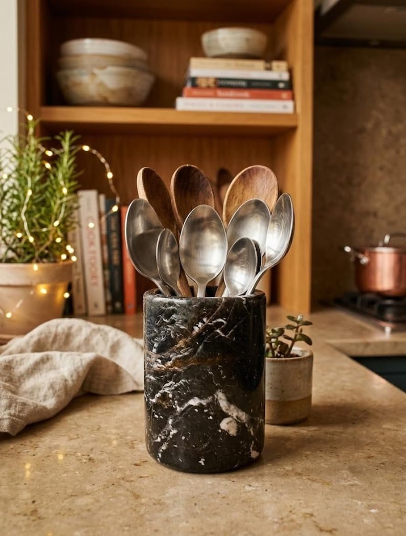 May include: A black and gold marble utensil holder filled with wooden and silver spoons. The cylindrical holder sits on a light-colored countertop. A small potted plant and a stack of books are in the background.