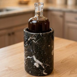 May include: A clear glass bottle filled with a dark liquid sits inside a black marble wine chiller. The chiller has white veining throughout. The bottle has a rounded base and a long, narrow neck. The background is blurred.