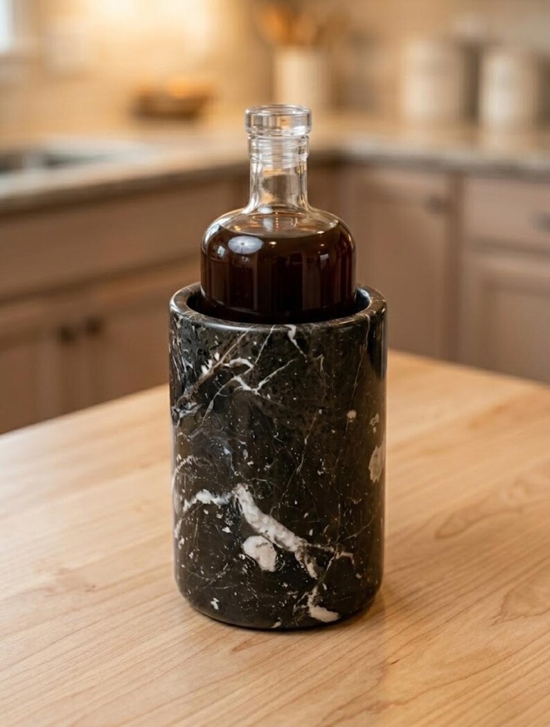 May include: A clear glass bottle filled with a dark liquid sits inside a black marble wine chiller. The chiller has white veining throughout. The bottle has a rounded base and a long, narrow neck. The background is blurred.