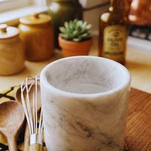 May include: A white marble kitchen utensil holder with gray veining. A wooden spoon and whisk rest on a patterned yellow and black cloth. Other kitchen items are in the background, including a bottle of olive oil and a small potted plant.