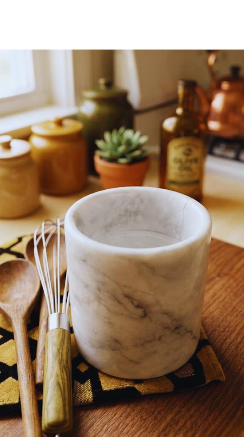 May include: A white marble kitchen utensil holder with gray veining. A wooden spoon and whisk rest on a patterned yellow and black cloth. Other kitchen items are in the background, including a bottle of olive oil and a small potted plant.