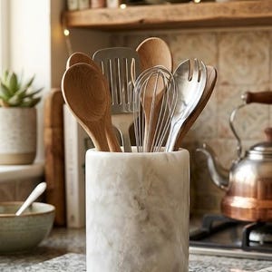 May include: A collection of kitchen utensils, including wooden spoons, a whisk, and a spatula, are displayed in a white marble utensil holder. The holder sits on a speckled countertop, with a kettle and a shelf of books in the background.
