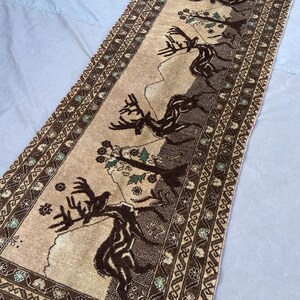 May include: A rectangular runner rug featuring a tan center field with dark brown silhouettes of deer and trees. The border displays a geometric pattern in dark brown and tan with small accents of light blue, finished with white fringe at the ends.