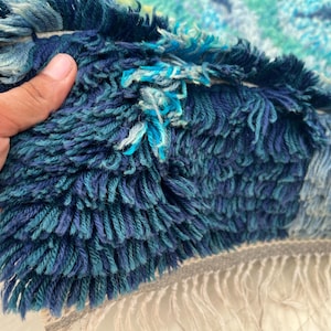 May include: Close-up of a textured rug with a mix of blue and teal hues. The rug has a shaggy, fringed texture with varying shades of blue, creating a layered effect. The fringe is a light gray color.
