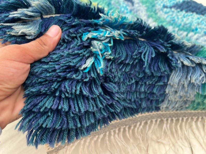 May include: Close-up of a textured rug with a mix of blue and teal hues. The rug has a shaggy, fringed texture with varying shades of blue, creating a layered effect. The fringe is a light gray color.