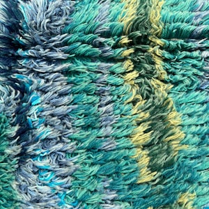 May include: Close-up of a textured rug with a shaggy pile. The rug features a blend of teal, blue, and yellow hues, creating a vibrant and tactile surface. The design has a striped pattern.