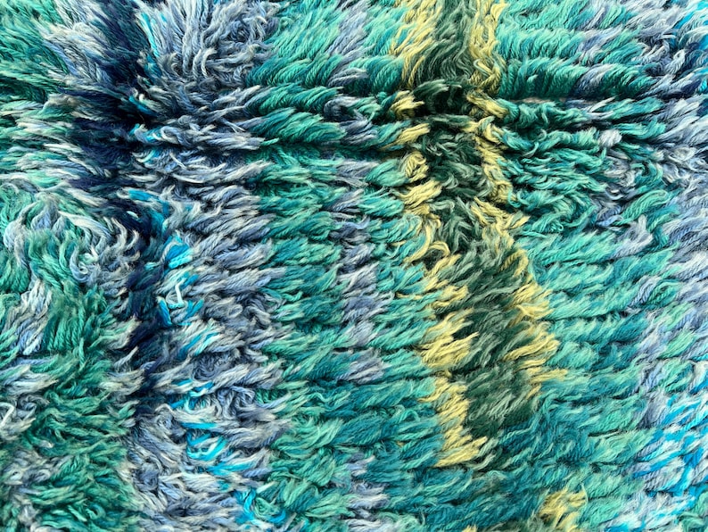 May include: Close-up of a textured rug with a shaggy pile. The rug features a blend of teal, blue, and yellow hues, creating a vibrant and tactile surface. The design has a striped pattern.