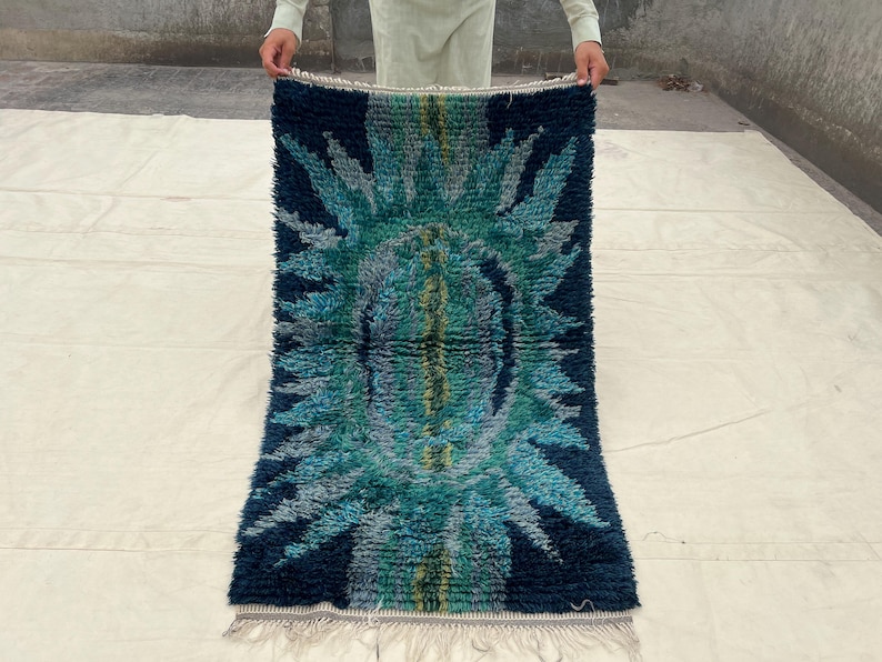 May include: A rectangular rug with a central design in shades of teal, blue, and green. The rug has a dark blue border and a fringe along the bottom edge. The design features a central oval shape with radiating patterns.