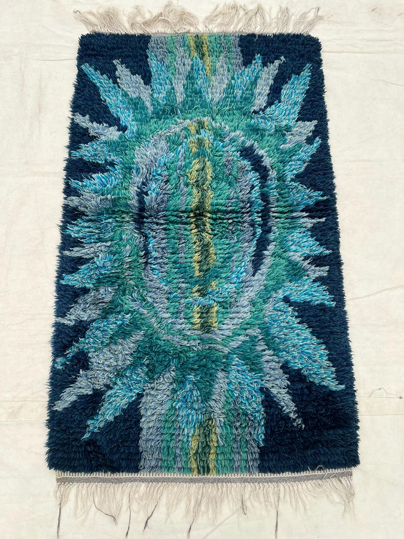 May include: A rectangular, shaggy rug with a vibrant, abstract design. The rug features a dark navy blue background with a central pattern in shades of teal, turquoise, and light blue. The edges have a fringe.