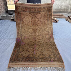 May include: A rectangular brown area rug featuring a repeating diamond pattern in darker brown tones. The rug has a decorative border with intricate detailing and a fringe along the top and bottom edges, with subtle hints of purple and orange accents.