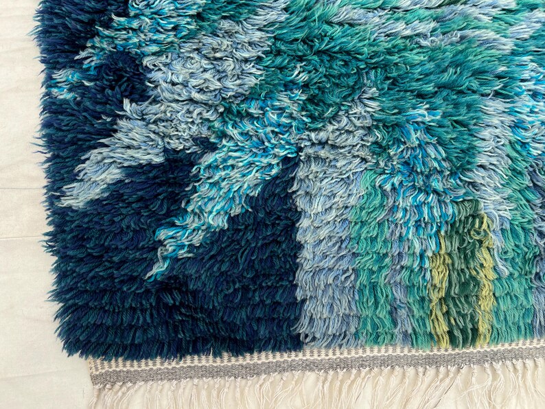 May include: A textured rug with a vibrant design. The rug features a mix of blues and greens, creating a layered effect. The edges are finished with a gray border and white fringe. The rug's design evokes a sense of nature.