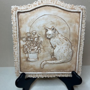 May include: A rectangular wall plaque featuring a raised relief design of a cat sitting beside a potted plant. The piece has a cream and light brown finish with a textured, ruffled border. The cat is positioned on the right, facing left.