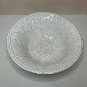 May include: A white ceramic serving bowl with a sunflower design. The bowl has a wide, shallow shape with a detailed embossed floral pattern. The rim features a decorative beaded edge, adding a touch of elegance to the piece.