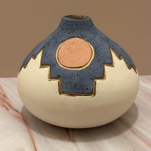May include: A ceramic vase with a rounded body and a narrow neck. The top half is a speckled blue, and the bottom half is cream-colored. A peach-colored circle is centered on the blue section, outlined in gold. Gold geometric shapes decorate the cream section.