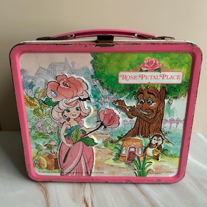 May include: Vintage pink metal lunchbox featuring a Rose Petal Place design. The artwork depicts a girl with a rose headdress, a tree playing a flute, and the words "Rose Petal Place". The lunchbox has a pink handle and trim.