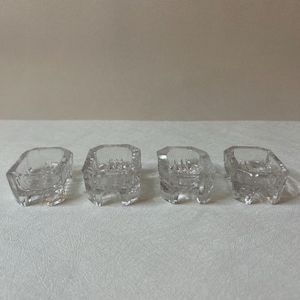 May include: A set of four small, clear glass knife rests, each featuring a rectangular, open-center design with four short, sturdy feet at the corners. The glass is transparent and smooth, with a slightly faceted appearance on the exterior edges.