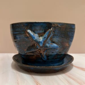 May include: A handcrafted ceramic planter with a blue glaze and a starfish design. The planter sits on a matching saucer. The dark blue glaze has lighter blue highlights, creating a textured, ocean-like appearance. A decorative piece for plants.