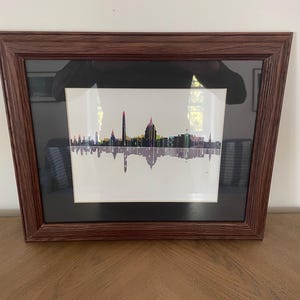 May include: Framed art print featuring a colorful skyline silhouette with a mirrored reflection. The artwork is set against a white background and framed in a dark brown wooden frame. The image is displayed on a wooden surface.