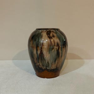 May include: A ceramic vase with a rounded body and a narrow opening, featuring a glossy glaze with blended streaks of dark brown, cream, and muted blue-green colors that drip vertically down the surface toward a solid brown base.
