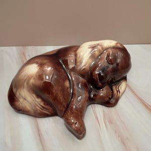May include: A ceramic figurine of a sleeping bear, featuring a glossy finish. The bear is primarily brown with lighter beige accents on its head and back. The figurine is curled up, with its paws tucked in.