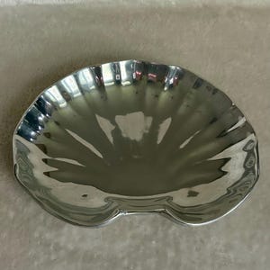 May include: A shiny, silver-colored seashell-shaped serving dish. The dish has a scalloped edge and a smooth, reflective surface. The dish has a double-lobed base. The dish is likely made of metal.