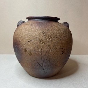 May include: A round, brown ceramic jar with a textured, matte finish and a dark rim. The vessel features two small, rounded handles on the sides and an etched design of slender, arching grass blades with small flower blossoms on the front surface.